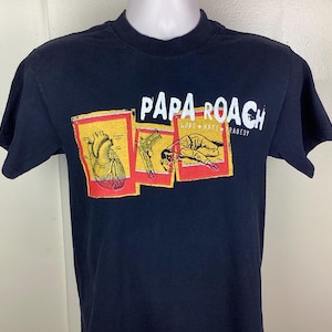 Puede incluir: Camiseta negra con el nombre de la banda "Papa Roach" en blanco, y las palabras "Love Hate Tragedy" debajo. La camiseta tiene tres gráficos: un corazón, una pistola y una persona tumbada. Los gráficos son en amarillo y rojo.