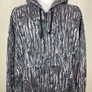 May include: A full-zip hooded sweatshirt featuring a Realtree camouflage pattern with bark, leaf, and branch motifs in shades of brown, gray, black, and muted green. The garment includes a front zipper, a hood with drawstrings, and dark cuffs.