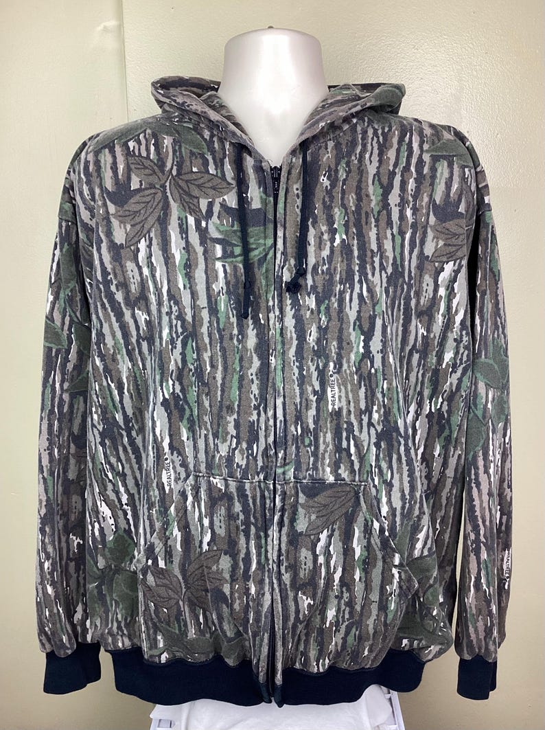 May include: A full-zip hooded sweatshirt featuring a Realtree camouflage pattern with bark, leaf, and branch motifs in shades of brown, gray, black, and muted green. The garment includes a front zipper, a hood with drawstrings, and dark cuffs.