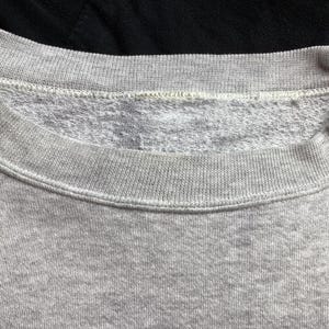 May include: Close-up of a gray sweatshirt collar. The collar is ribbed and the inside is a lighter gray, textured material. The sweatshirt is a light gray color and appears to be made of a soft fabric.
