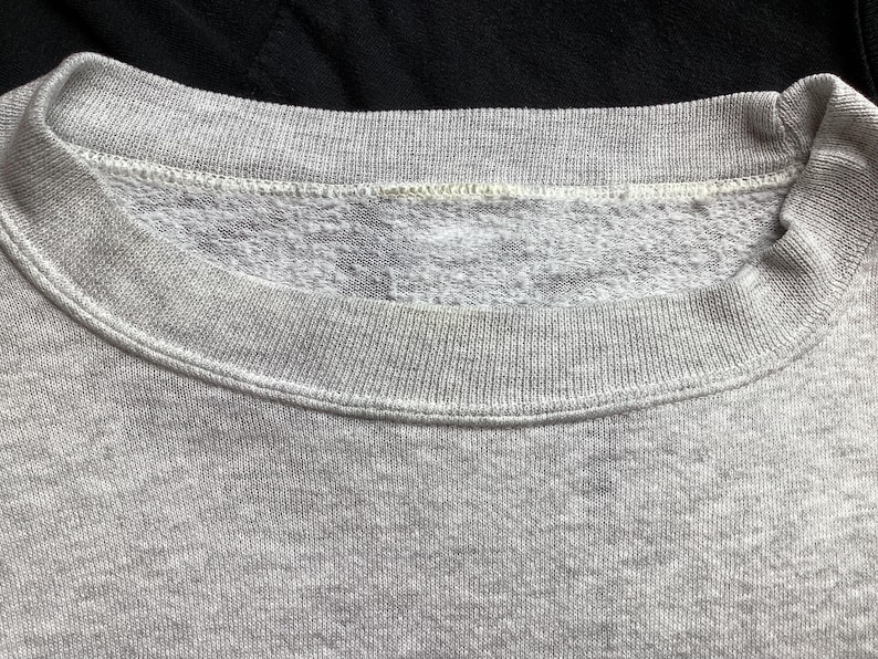 May include: Close-up of a gray sweatshirt collar. The collar is ribbed and the inside is a lighter gray, textured material. The sweatshirt is a light gray color and appears to be made of a soft fabric.