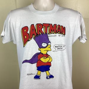 May include: White t-shirt with a cartoon image of Bart Simpson dressed as a superhero. The text "Bartman Avenger of Evil" is above the image and "Watch it, Dude." is below the image.