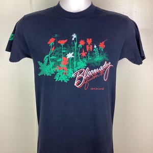 Pu&ograve; includere: T-shirt blu navy con un design floreale con accenti rossi, bianchi e verdi. Il testo "Bloomsday Finisher '85 Spokane" &egrave; stampato. La t-shirt &egrave; esposta su un manichino.