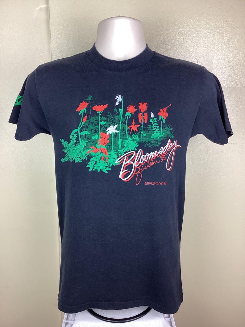 Pu&ograve; includere: T-shirt blu navy con un design floreale con accenti rossi, bianchi e verdi. Il testo "Bloomsday Finisher '85 Spokane" &egrave; stampato. La t-shirt &egrave; esposta su un manichino.