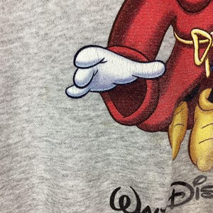 May include: Close-up of a gray sweatshirt featuring a cartoon character in a red outfit with a white glove. The Walt Disney logo is printed in black at the bottom right corner of the image.
