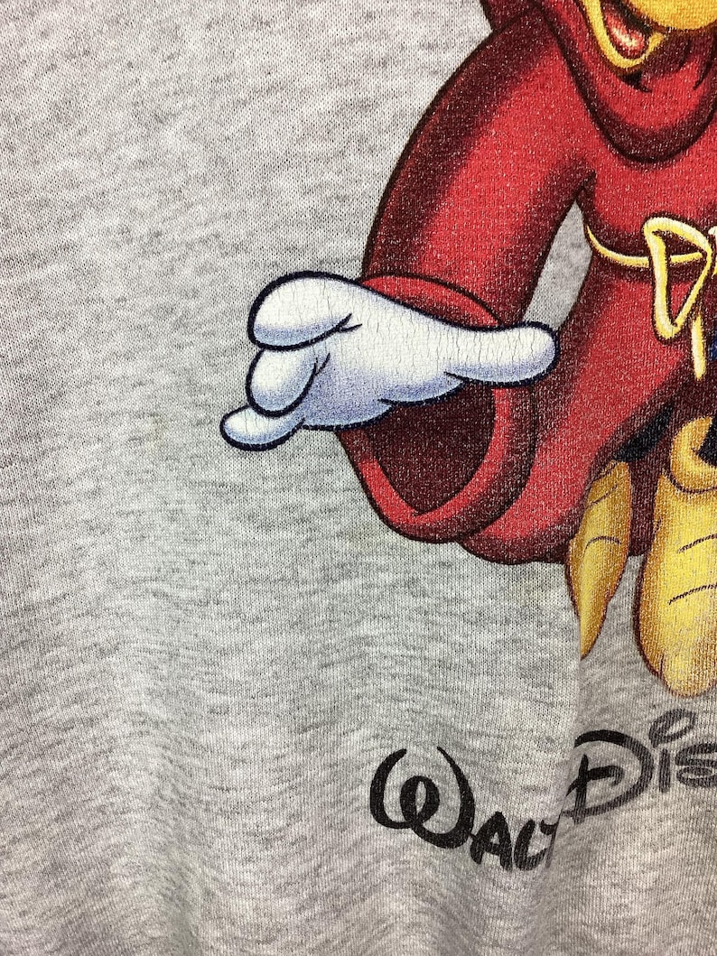 May include: Close-up of a gray sweatshirt featuring a cartoon character in a red outfit with a white glove. The Walt Disney logo is printed in black at the bottom right corner of the image.