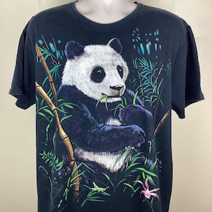 May include: Black t-shirt with a panda bear illustration. The panda, in black, white, and blue, is eating bamboo. Green bamboo stalks and leaves surround the panda, with a pink flower. The t-shirt is on a mannequin.