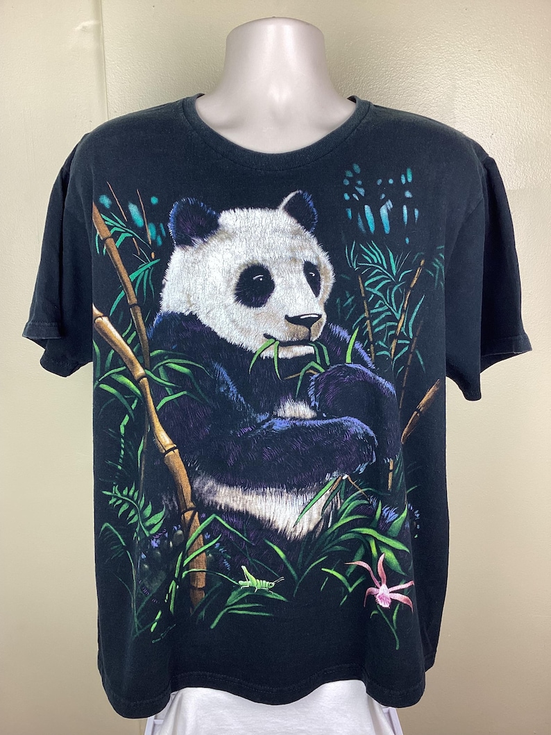 May include: Black t-shirt with a panda bear illustration. The panda, in black, white, and blue, is eating bamboo. Green bamboo stalks and leaves surround the panda, with a pink flower. The t-shirt is on a mannequin.