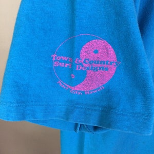 K&ouml;nnte beinhalten: Ein t&uuml;rkisblaues T-Shirt mit einem rosafarbenen Kreislogo, auf dem "Town & Country Surf Designs" und "Pearl City, Hawaii" steht.