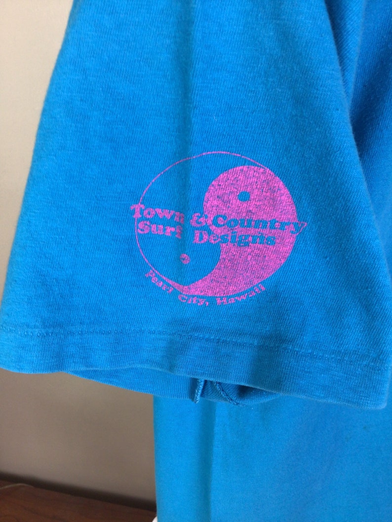 K&ouml;nnte beinhalten: Ein t&uuml;rkisblaues T-Shirt mit einem rosafarbenen Kreislogo, auf dem "Town & Country Surf Designs" und "Pearl City, Hawaii" steht.