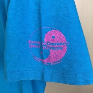 K&ouml;nnte beinhalten: Ein t&uuml;rkisfarbenes T-Shirt mit einem rosafarbenen Yin-Yang-Symbol und dem Text "Town & Country Surf Designs Pearl City, Hawaii" auf dem &Auml;rmel.