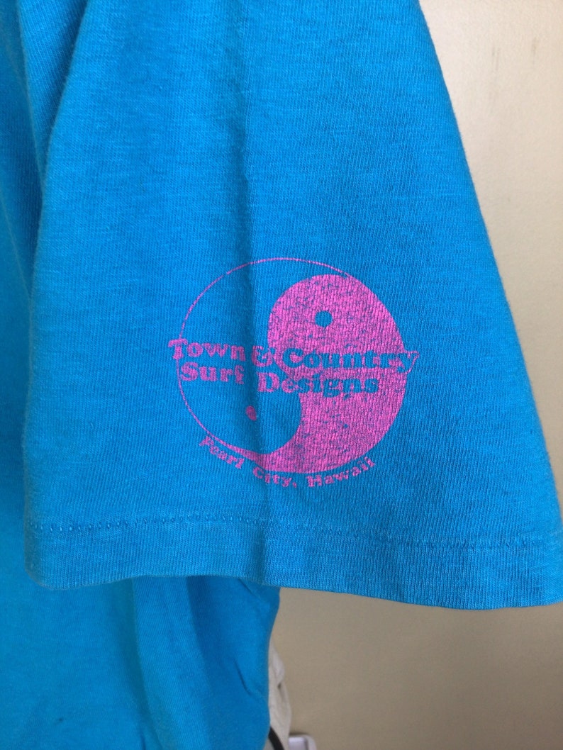 K&ouml;nnte beinhalten: Ein t&uuml;rkisfarbenes T-Shirt mit einem rosafarbenen Yin-Yang-Symbol und dem Text "Town & Country Surf Designs Pearl City, Hawaii" auf dem &Auml;rmel.