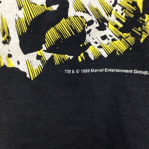 Pu&ograve; includere: Maglietta nera con una grafica in stile fumetto giallo e bianco. Il testo "TM & &copy; 1989 Marvel Entertainment Group" &egrave; stampato sul fondo della maglietta.