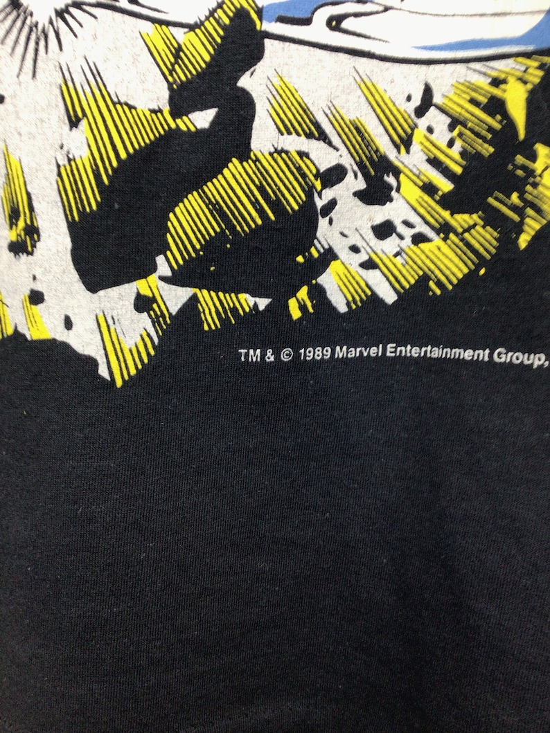 Pu&ograve; includere: Maglietta nera con una grafica in stile fumetto giallo e bianco. Il testo "TM & &copy; 1989 Marvel Entertainment Group" &egrave; stampato sul fondo della maglietta.