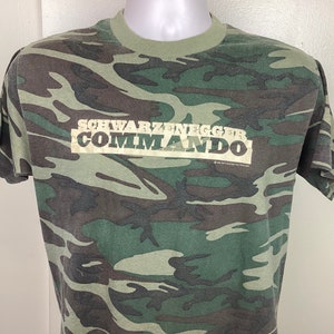 Può includere: Una t-shirt mimetica verde, marrone e bianca con la scritta "Schwarzenegger Commando" stampata sul davanti.