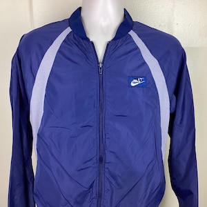 May include: A blue and white windbreaker jacket with a zip fastening and a Nike logo on the left chest. The jacket has white panels on the sleeves and back.