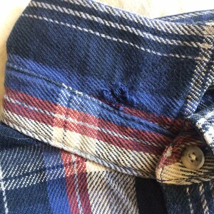 May include: A blue, red and beige plaid flannel shirt with a button fastening. The shirt has a small tear in the collar.
