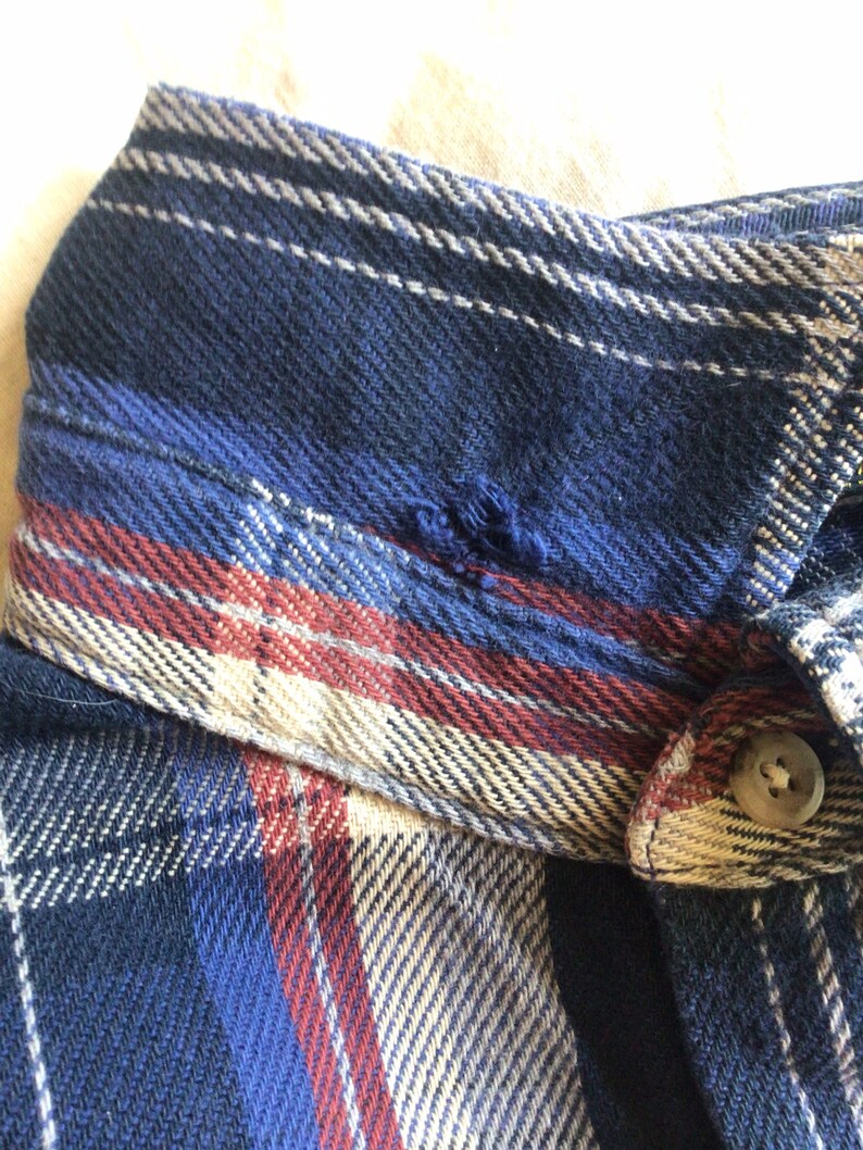 May include: A blue, red and beige plaid flannel shirt with a button fastening. The shirt has a small tear in the collar.