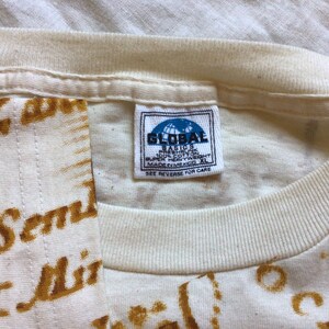 May include: White t-shirt with a brown print of the words "I Send" and "I Miss" repeated across the fabric. The shirt has a white tag with the text "Global Basics" and "Made in Bangladesh" on it.