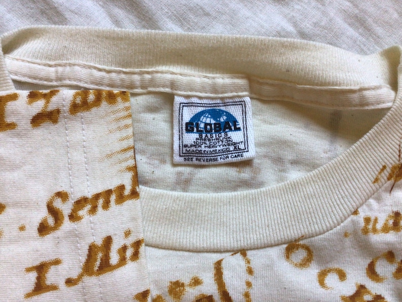 May include: White t-shirt with a brown print of the words "I Send" and "I Miss" repeated across the fabric. The shirt has a white tag with the text "Global Basics" and "Made in Bangladesh" on it.