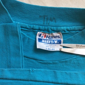 K&ouml;nnte beinhalten: Ein t&uuml;rkisblaues Hanes Beefeater T-Shirt-Etikett mit wei&szlig;em Text. Das Etikett lautet "100% Baumwolle Hergestellt in den USA L 42-44".