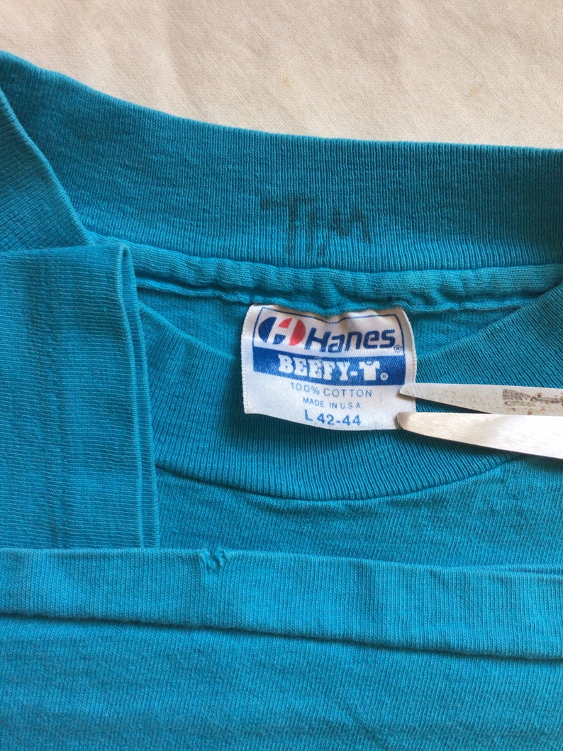 K&ouml;nnte beinhalten: Ein t&uuml;rkisblaues Hanes Beefeater T-Shirt-Etikett mit wei&szlig;em Text. Das Etikett lautet "100% Baumwolle Hergestellt in den USA L 42-44".