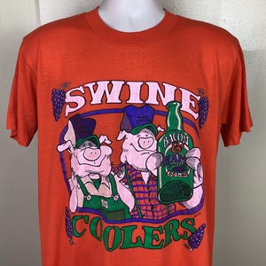 Op de afbeelding: Oranje T-shirt met een afbeelding van twee cartoon varkens die een fles "Bacon & Ham Swine Cooler" wijn vasthouden. De tekst "Swine Coolers" is gedrukt in groene en paarse letters.