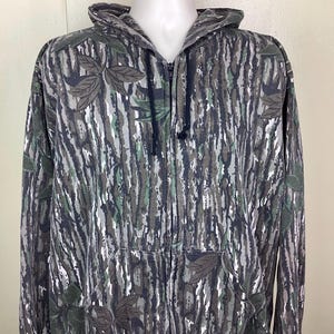 May include: A zip-up hooded sweatshirt featuring a Realtree camouflage pattern in shades of gray, black, and green. The garment includes a full-length front zipper, a drawstring hood with black cords, and two front pockets integrated into the design.