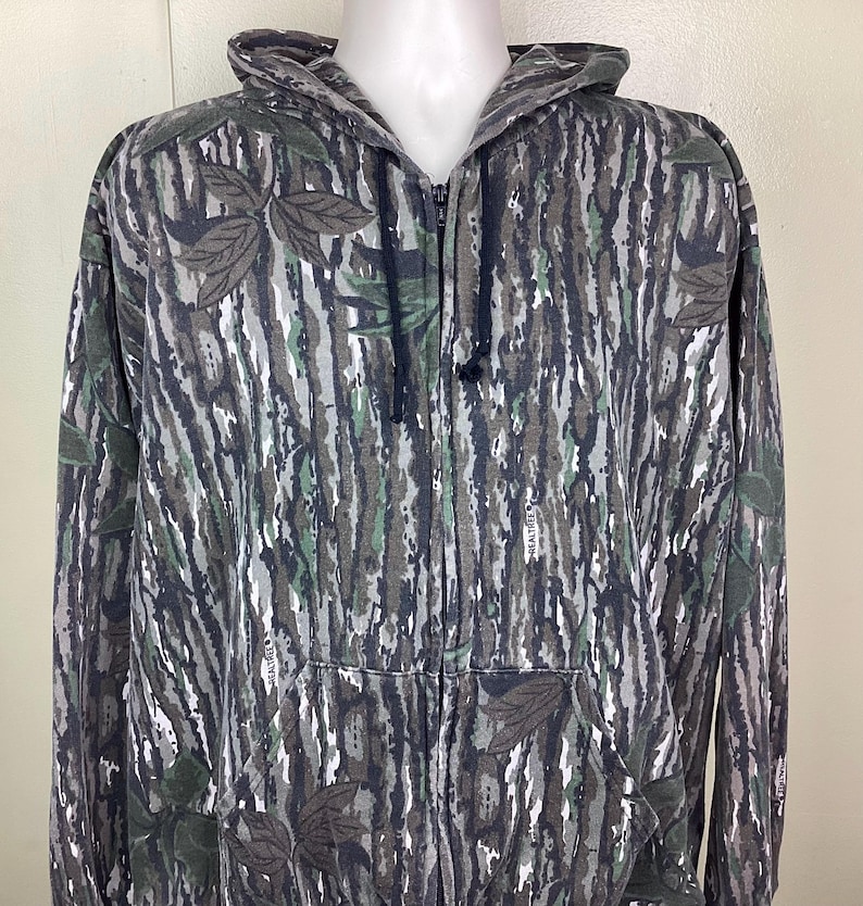 May include: A zip-up hooded sweatshirt featuring a Realtree camouflage pattern in shades of gray, black, and green. The garment includes a full-length front zipper, a drawstring hood with black cords, and two front pockets integrated into the design.