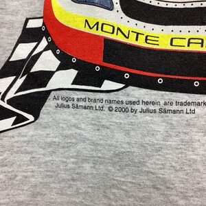 May include: A gray t-shirt with a black and white checkered pattern and a red, yellow, and black illustration of a Monte Carlo car. The text "Monte Carlo" is visible on the car. The text "All logos and brand names used herein are trademark Julius S&auml;mann Ltd. &copy; 2000 by Julius S&auml;mann Ltd" is printed on the bottom of the shirt.