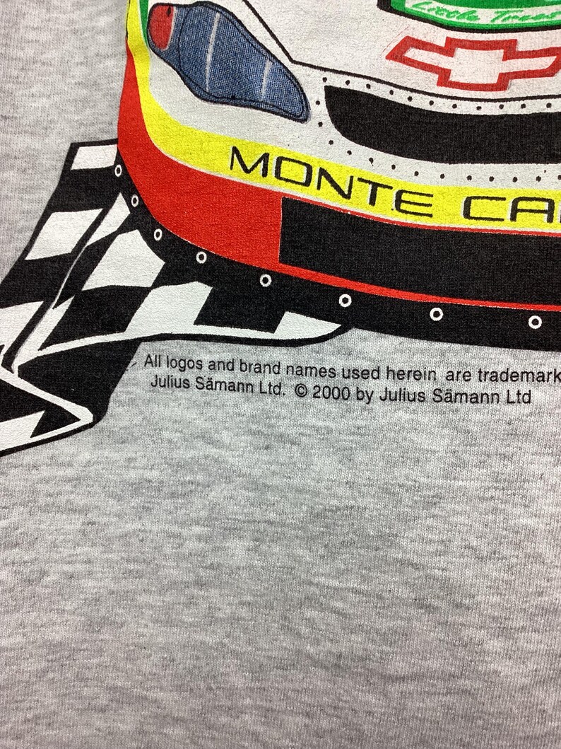 May include: A gray t-shirt with a black and white checkered pattern and a red, yellow, and black illustration of a Monte Carlo car. The text "Monte Carlo" is visible on the car. The text "All logos and brand names used herein are trademark Julius S&auml;mann Ltd. &copy; 2000 by Julius S&auml;mann Ltd" is printed on the bottom of the shirt.
