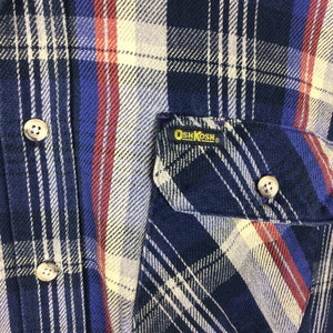 May include: A blue, red and white plaid flannel shirt with a patch pocket on the left side. The pocket has a yellow OshKosh label.