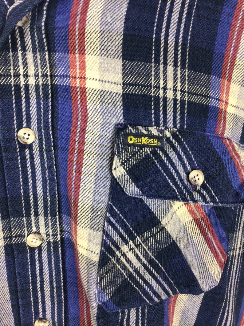 May include: A blue, red and white plaid flannel shirt with a patch pocket on the left side. The pocket has a yellow OshKosh label.