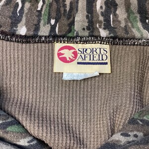 May include: A brown ribbed fabric garment featuring a camouflage pattern in shades of green, black, and brown. A rectangular white tag is sewn onto the interior, displaying a red circular logo with a white eagle and the text SPORTS AFIELD.