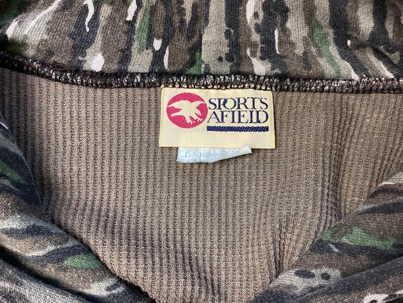 May include: A brown ribbed fabric garment featuring a camouflage pattern in shades of green, black, and brown. A rectangular white tag is sewn onto the interior, displaying a red circular logo with a white eagle and the text SPORTS AFIELD.