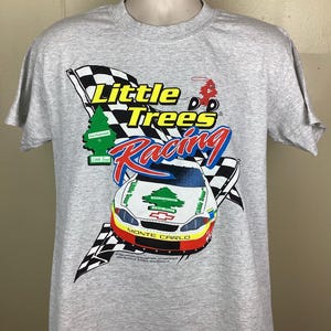 May include: A gray t-shirt with a graphic of a race car and the text "Little Trees Racing" in yellow and blue. The car is a white Chevrolet Monte Carlo with a green pine tree logo on the side. The car is driving towards a checkered flag.