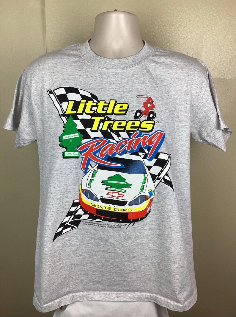May include: A gray t-shirt with a graphic of a race car and the text "Little Trees Racing" in yellow and blue. The car is a white Chevrolet Monte Carlo with a green pine tree logo on the side. The car is driving towards a checkered flag.