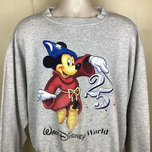 May include: A light gray sweatshirt featuring a graphic of Mickey Mouse dressed as a sorcerer, with a blue hat and red robe. The number "25" is next to Mickey, with the words "Walt Disney World" below.