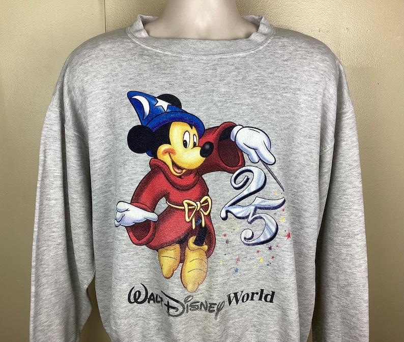 May include: A light gray sweatshirt featuring a graphic of Mickey Mouse dressed as a sorcerer, with a blue hat and red robe. The number "25" is next to Mickey, with the words "Walt Disney World" below.