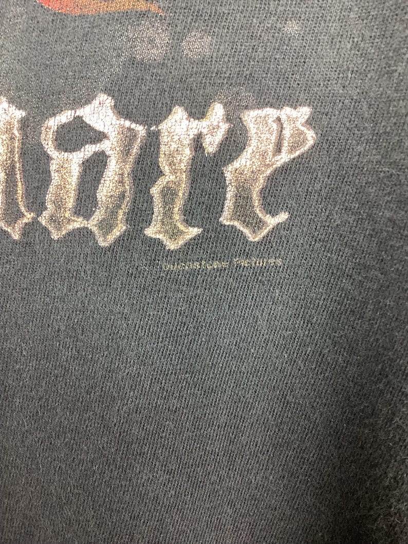 May include: Close-up of a dark gray t-shirt with the word "LIBER" in a stylized, metallic font. The text is slightly distressed, suggesting a vintage or worn appearance. The fabric has a subtle texture.