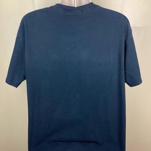 May include: A dark blue short-sleeved t-shirt displayed on a mannequin. The shirt has a crew neck and a slightly faded appearance. The fabric appears to be a soft cotton blend. The shirt is a solid color with no visible graphics or patterns.