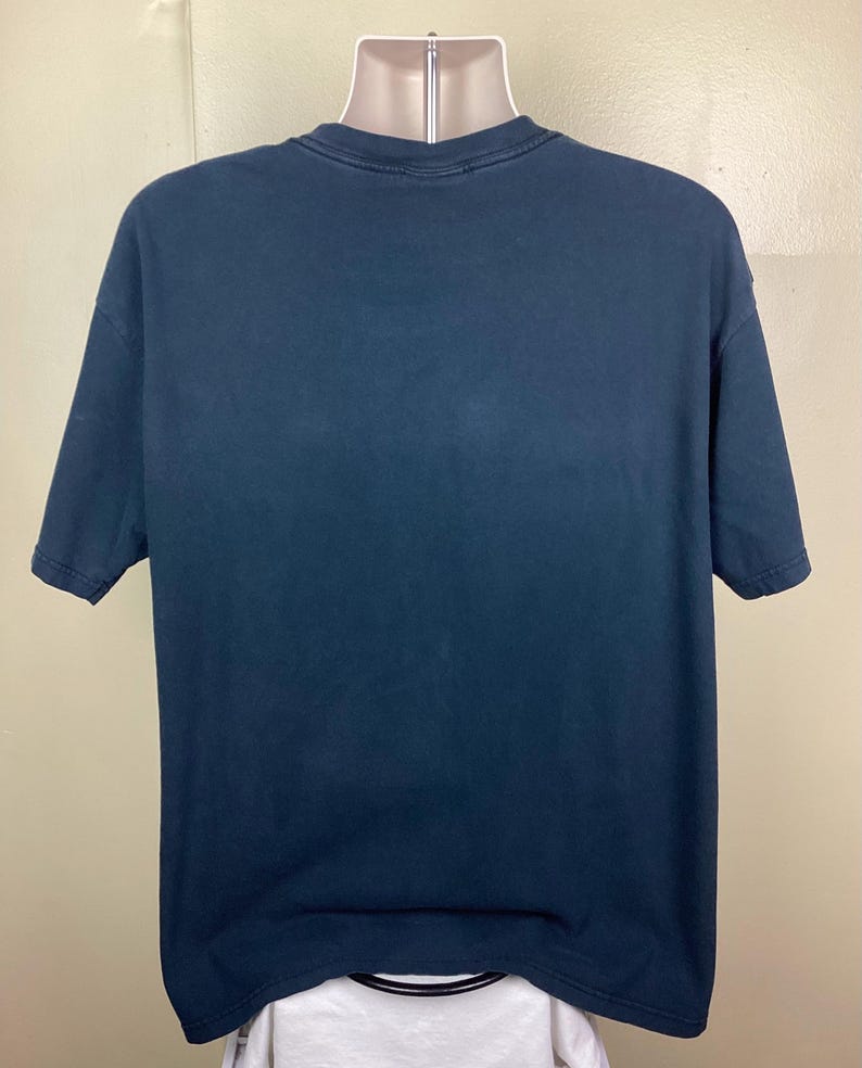 May include: A dark blue short-sleeved t-shirt displayed on a mannequin. The shirt has a crew neck and a slightly faded appearance. The fabric appears to be a soft cotton blend. The shirt is a solid color with no visible graphics or patterns.