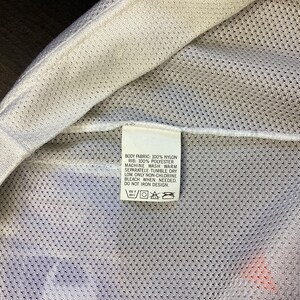 May include: Close-up of a white garment tag with care instructions. The tag is rectangular and features text detailing fabric composition and washing instructions. The garment appears to be made of nylon and polyester.