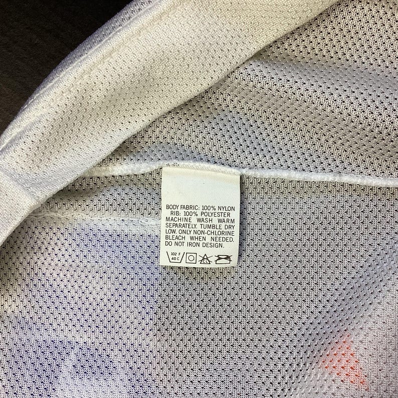 May include: Close-up of a white garment tag with care instructions. The tag is rectangular and features text detailing fabric composition and washing instructions. The garment appears to be made of nylon and polyester.