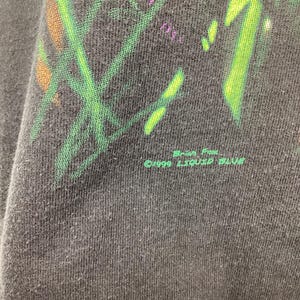May include: Close-up of a black t-shirt with a colourful graphic design featuring green, purple, and orange elements. The text "&copy;1999 LIQUID BLUE" is printed in green at the bottom of the shirt.