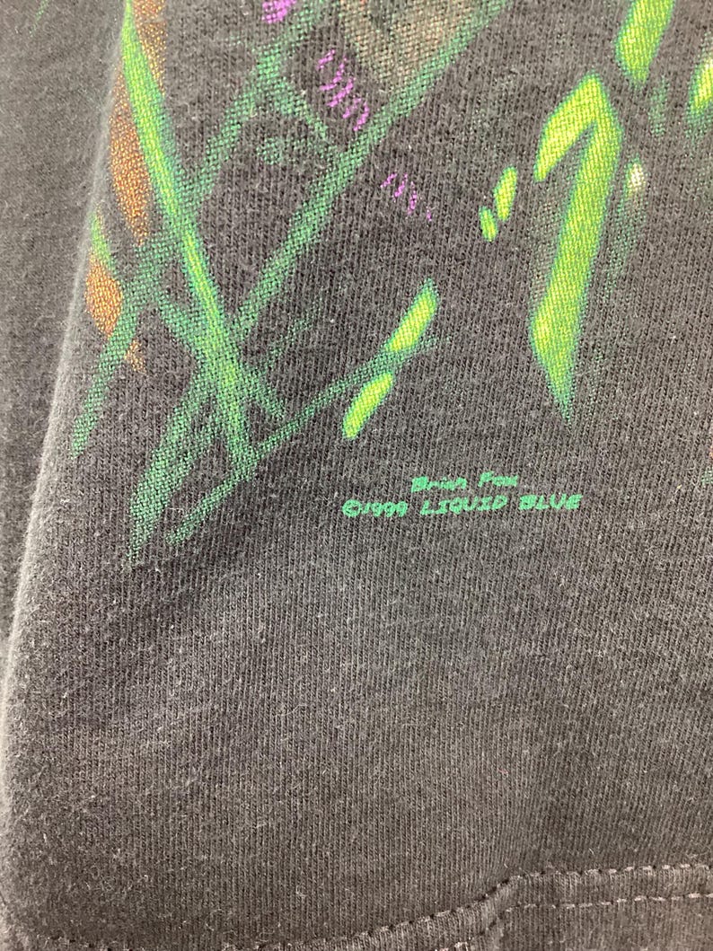 May include: Close-up of a black t-shirt with a colourful graphic design featuring green, purple, and orange elements. The text "&copy;1999 LIQUID BLUE" is printed in green at the bottom of the shirt.
