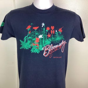 Pu&ograve; includere: T-shirt blu navy con un design floreale di fiori rossi, bianchi e verdi. Il testo "Bloomsday Finisher '85 Spokane" &egrave; stampato in bianco e rosso in corsivo. La maglietta ha maniche corte.