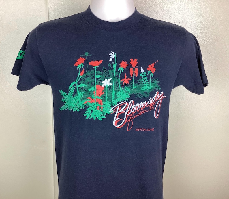 Pu&ograve; includere: T-shirt blu navy con un design floreale di fiori rossi, bianchi e verdi. Il testo "Bloomsday Finisher '85 Spokane" &egrave; stampato in bianco e rosso in corsivo. La maglietta ha maniche corte.