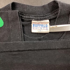 Pu&ograve; includere: T-shirt nero con un disegno verde sulla spalla. La maglietta ha un'etichetta con la scritta "BUFFALO SHIRTS" e la taglia "M". Un paio di forbici &egrave; posto sulla maglietta.
