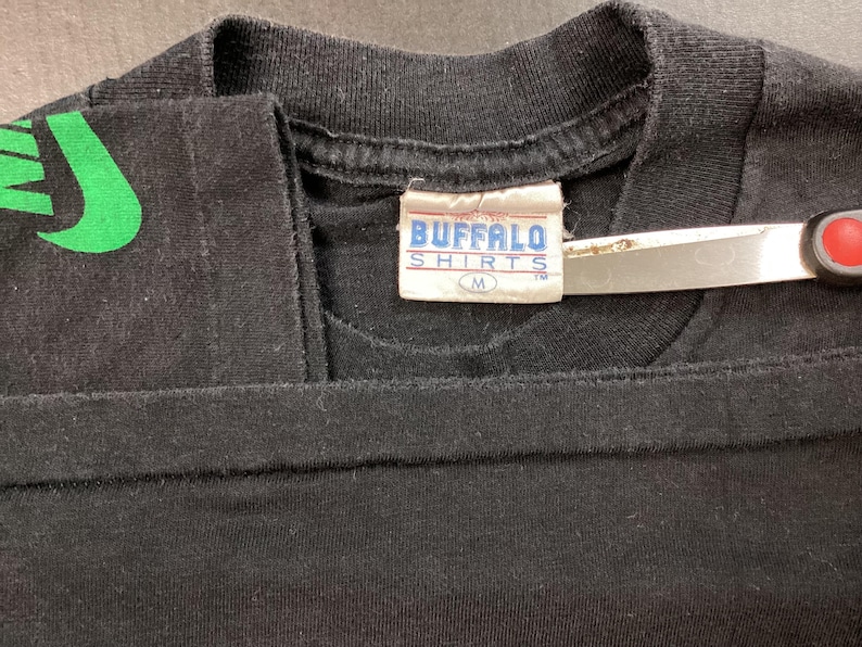 Pu&ograve; includere: T-shirt nero con un disegno verde sulla spalla. La maglietta ha un'etichetta con la scritta "BUFFALO SHIRTS" e la taglia "M". Un paio di forbici &egrave; posto sulla maglietta.